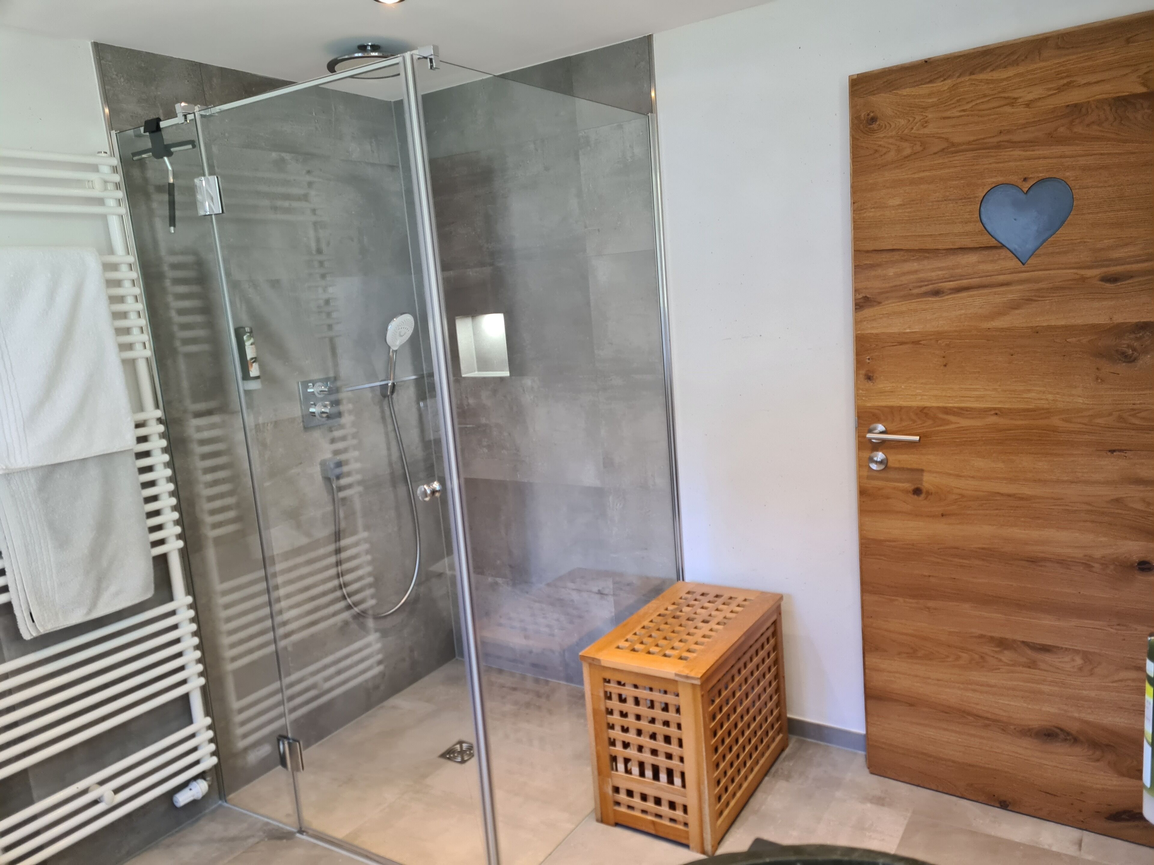 Un baño moderno con ducha de vidrio, una puerta de madera con un recorte en forma de corazón y un pequeño taburete de madera.