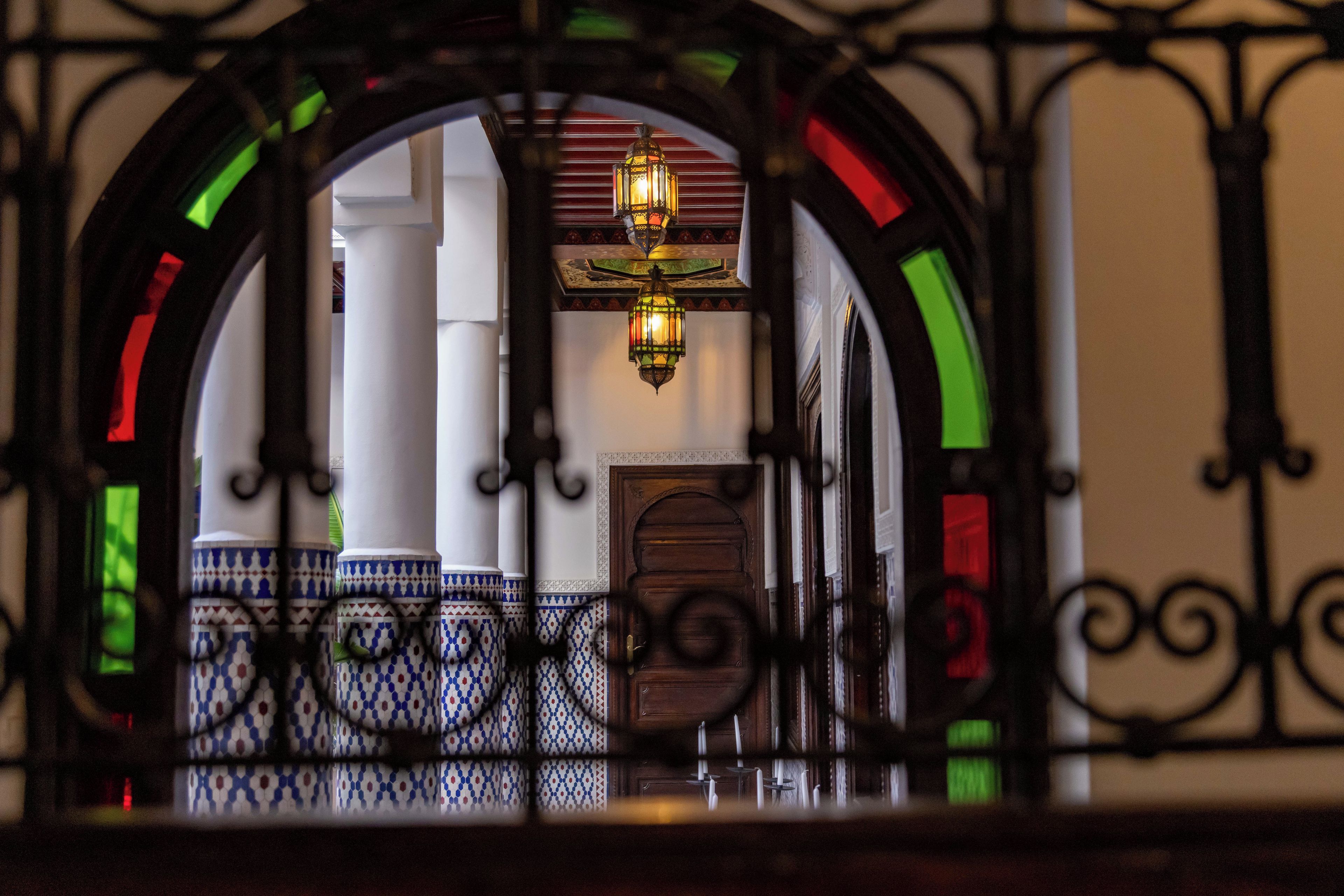A view through an ornate iron gate into a room with a patterned floor, a wooden door, and a hanging lantern.