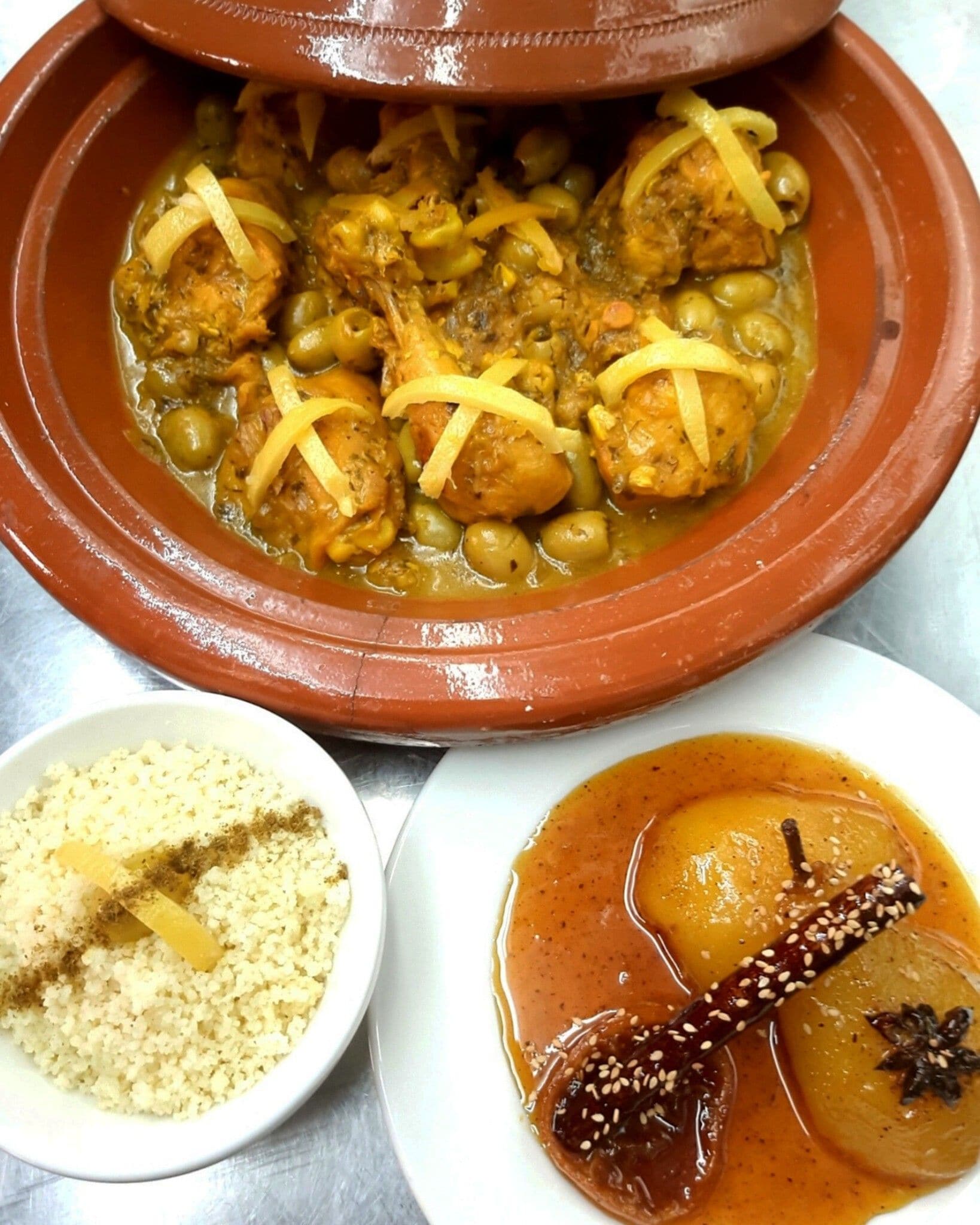 A traditional clay pot dish with chicken and olives, accompanied by a bowl of couscous and a bowl of stew with a cinnamon stick.