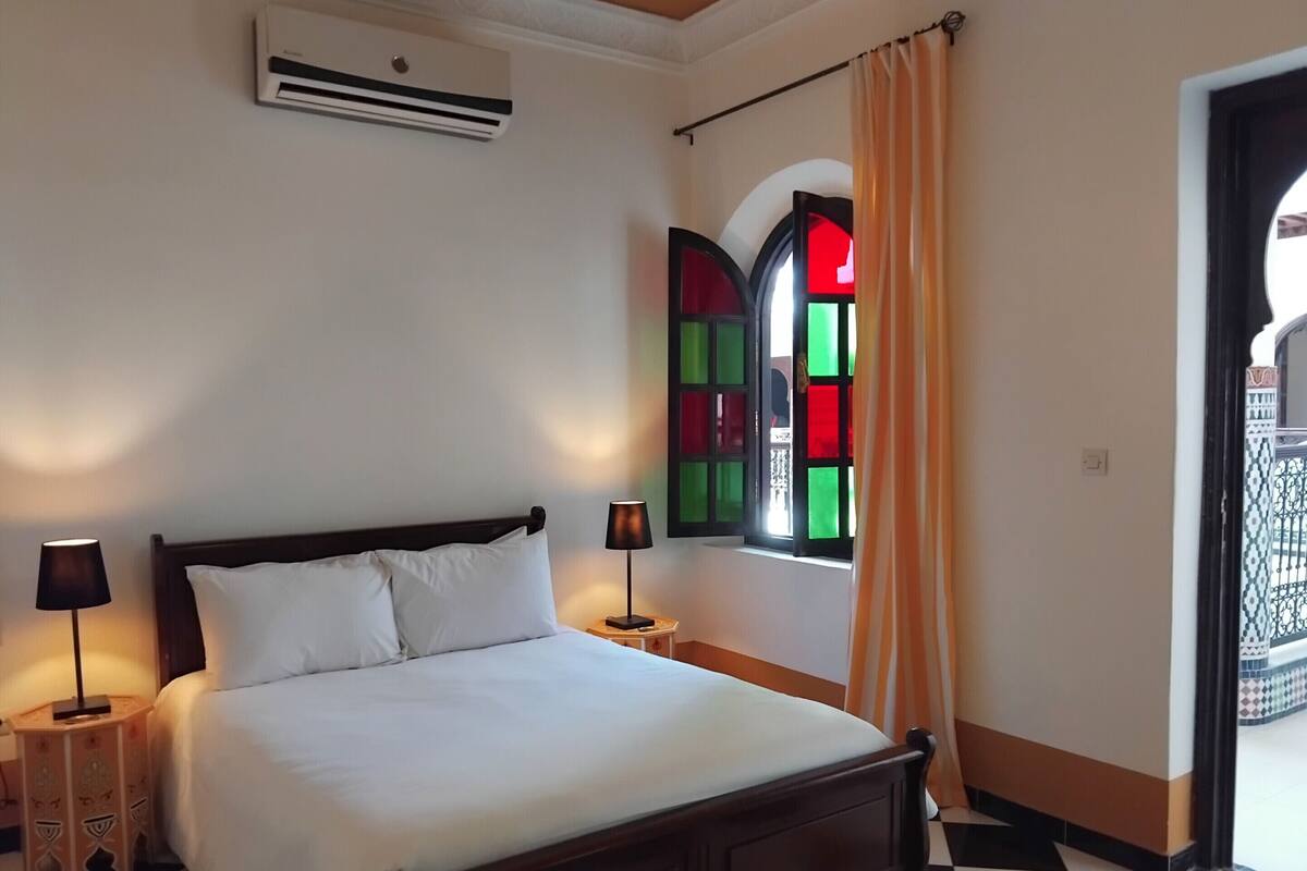 A hotel room with a bed, two bedside lamps, an air conditioning unit, a window with colorful stained glass, and a checkered floor.