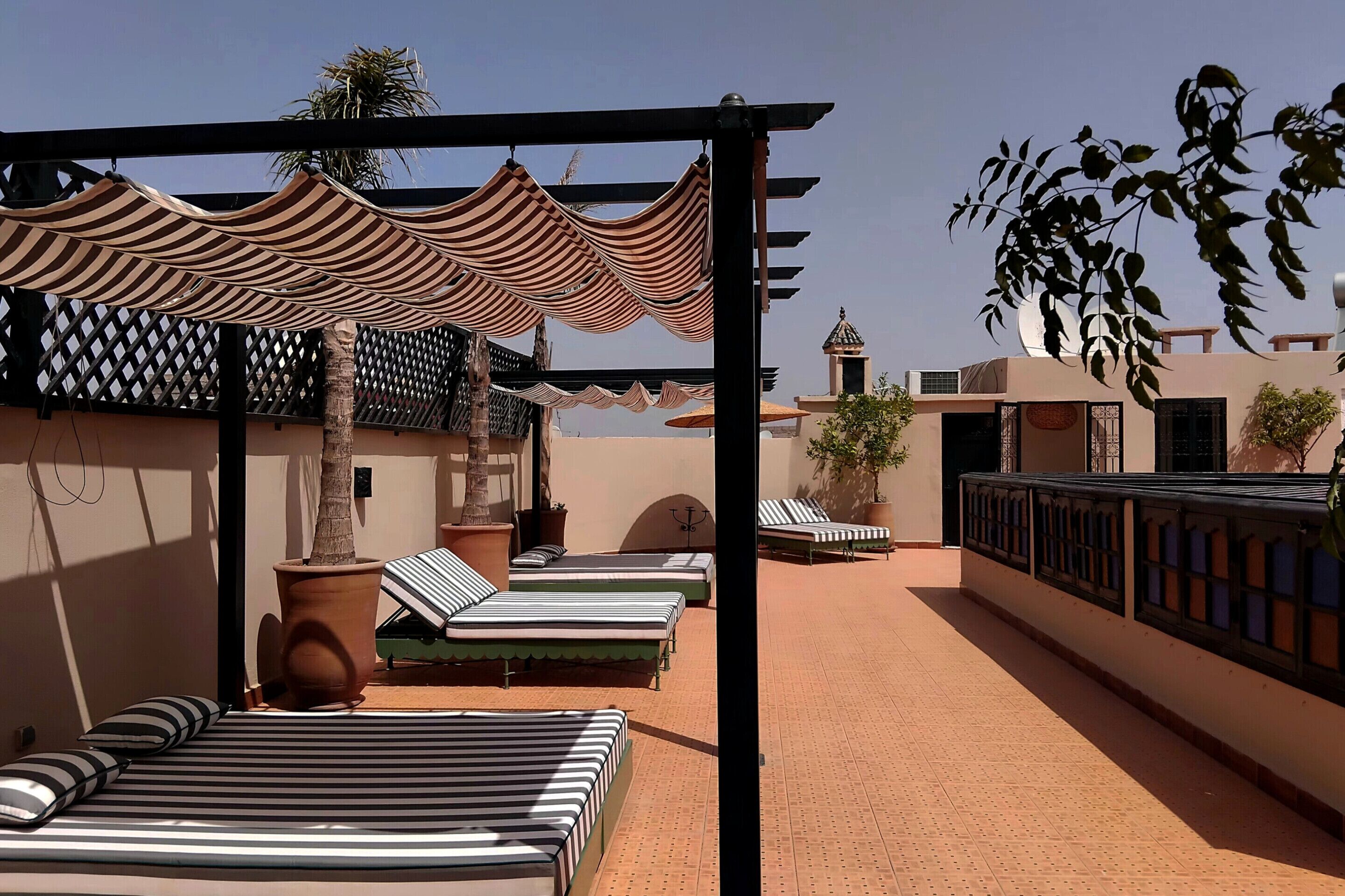 A rooftop terrace with striped lounge beds, a pergola providing shade, and a view of the surrounding buildings.