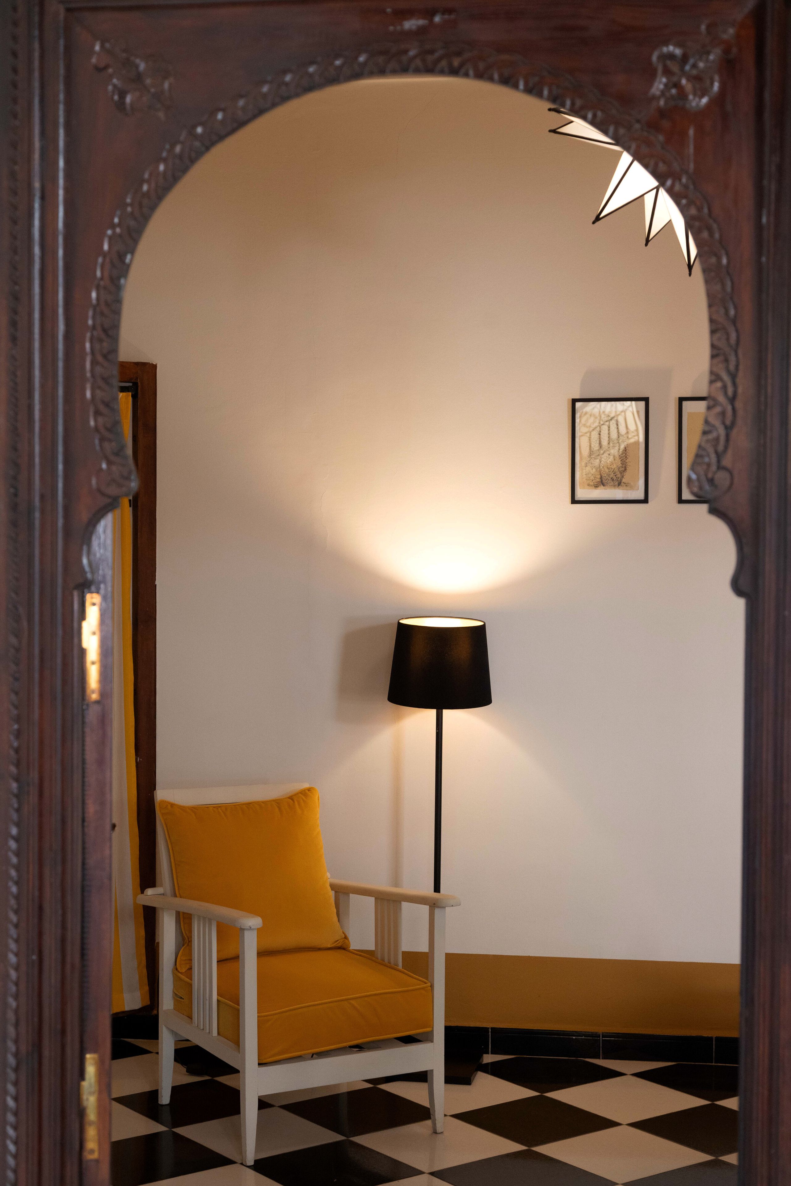 A room with a checkered floor, a white chair with a yellow cushion, a black floor lamp, and two framed pictures on the wall.