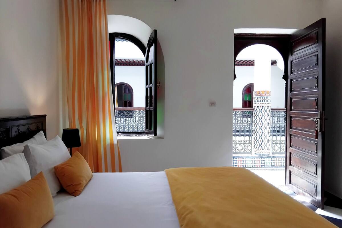 A hotel room with a bed, a window with curtains, and an open door leading to a balcony.
