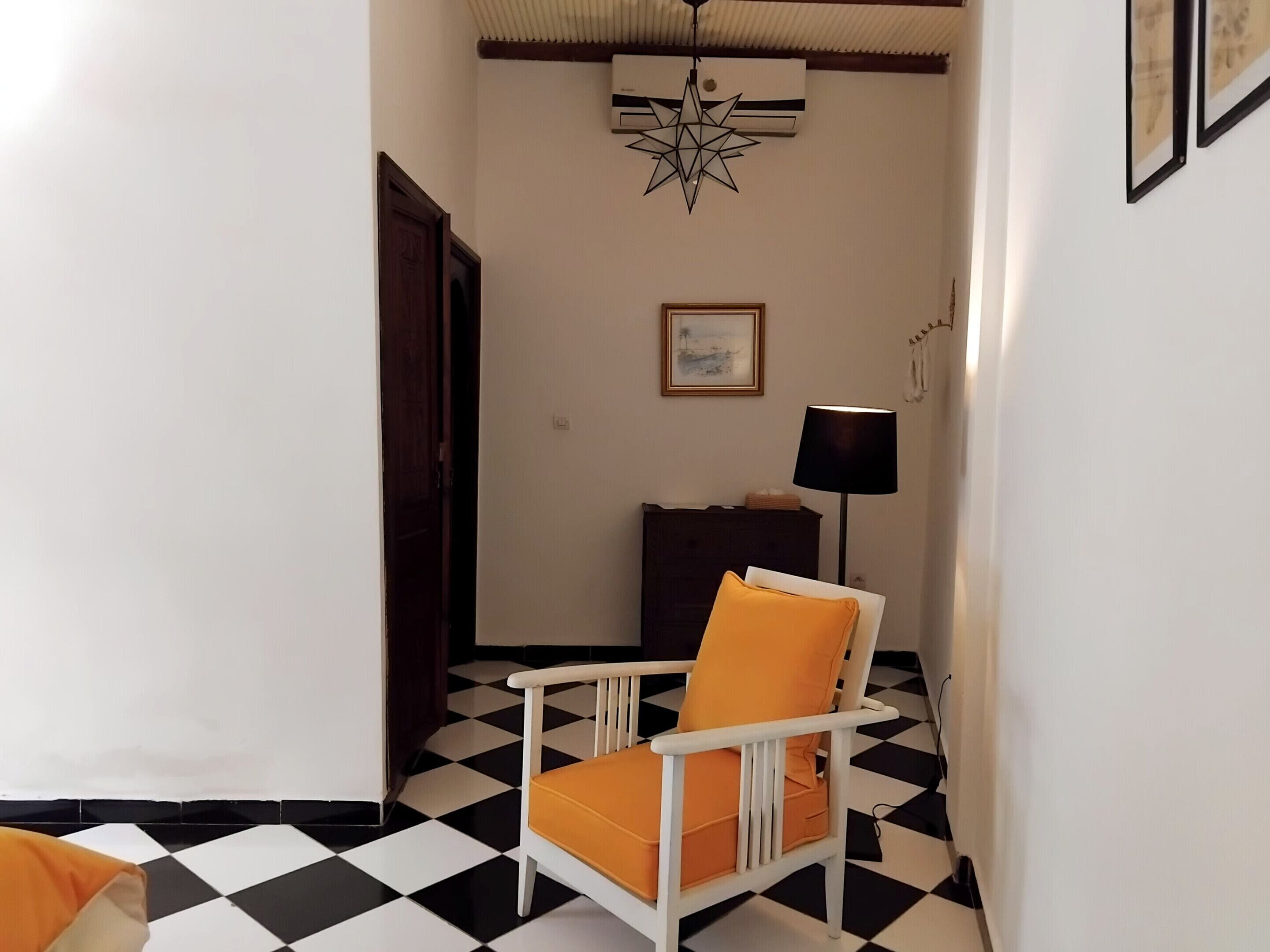 A hallway with a checkered floor, a white chair with an orange cushion, a dark wooden cabinet, and a framed picture on the wall.