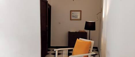 A hallway with a checkered floor, a white chair with an orange cushion, a dark wooden cabinet, and a framed picture on the wall.