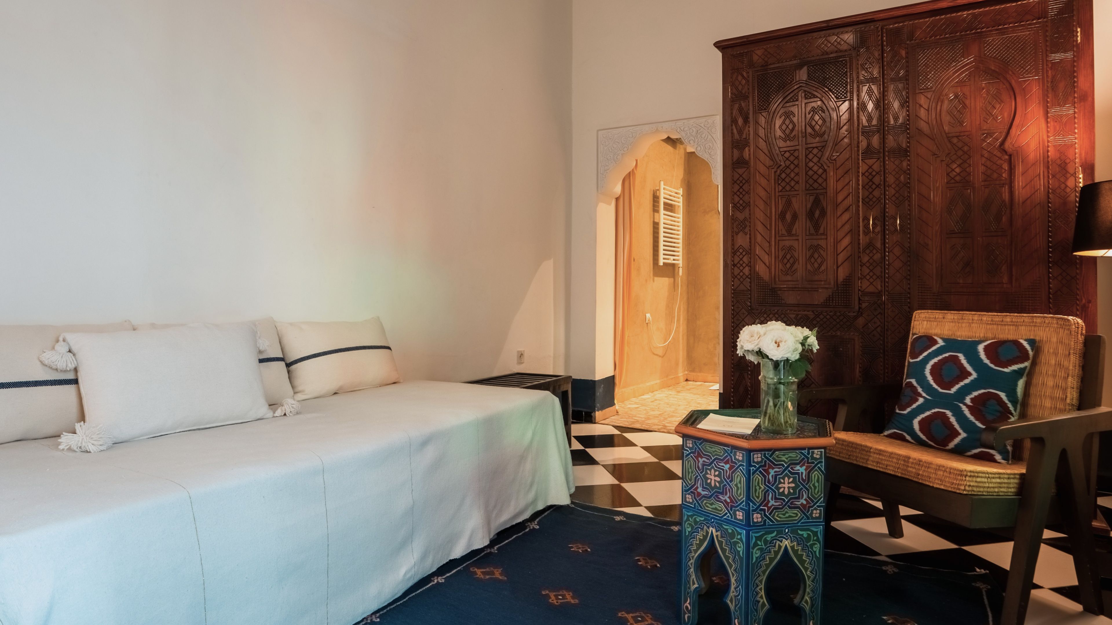 A room with a bed, a chair, a small table with a vase, and a wooden cabinet.