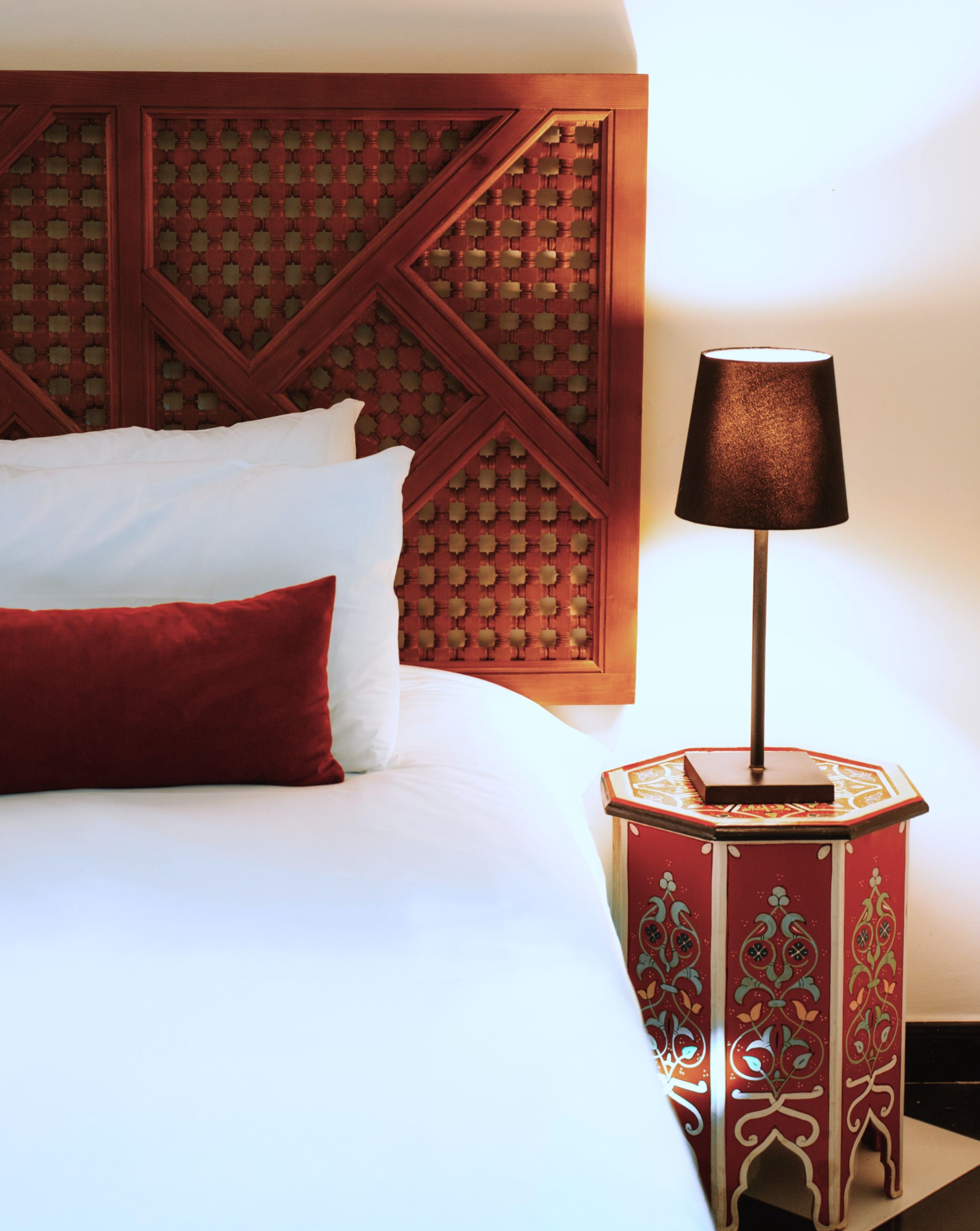 Een net opgemaakt bed met wit en rode beddengoed, een houten hoofdbord met een geometrisch patroon, en een klein tafeltje met een lamp ernaast.