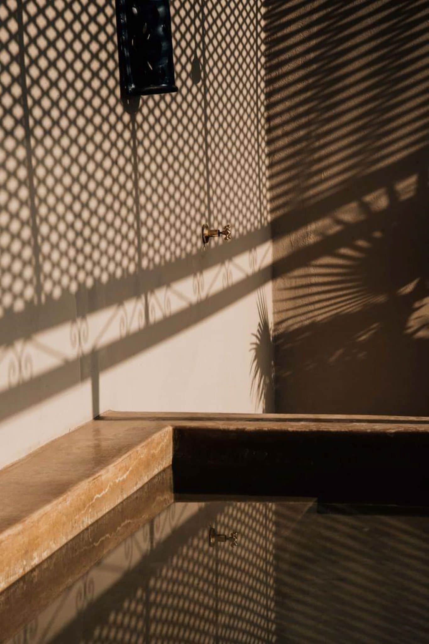 A wooden bench with a metal railing, a wall with a geometric pattern, and a shadow cast by a palm tree.