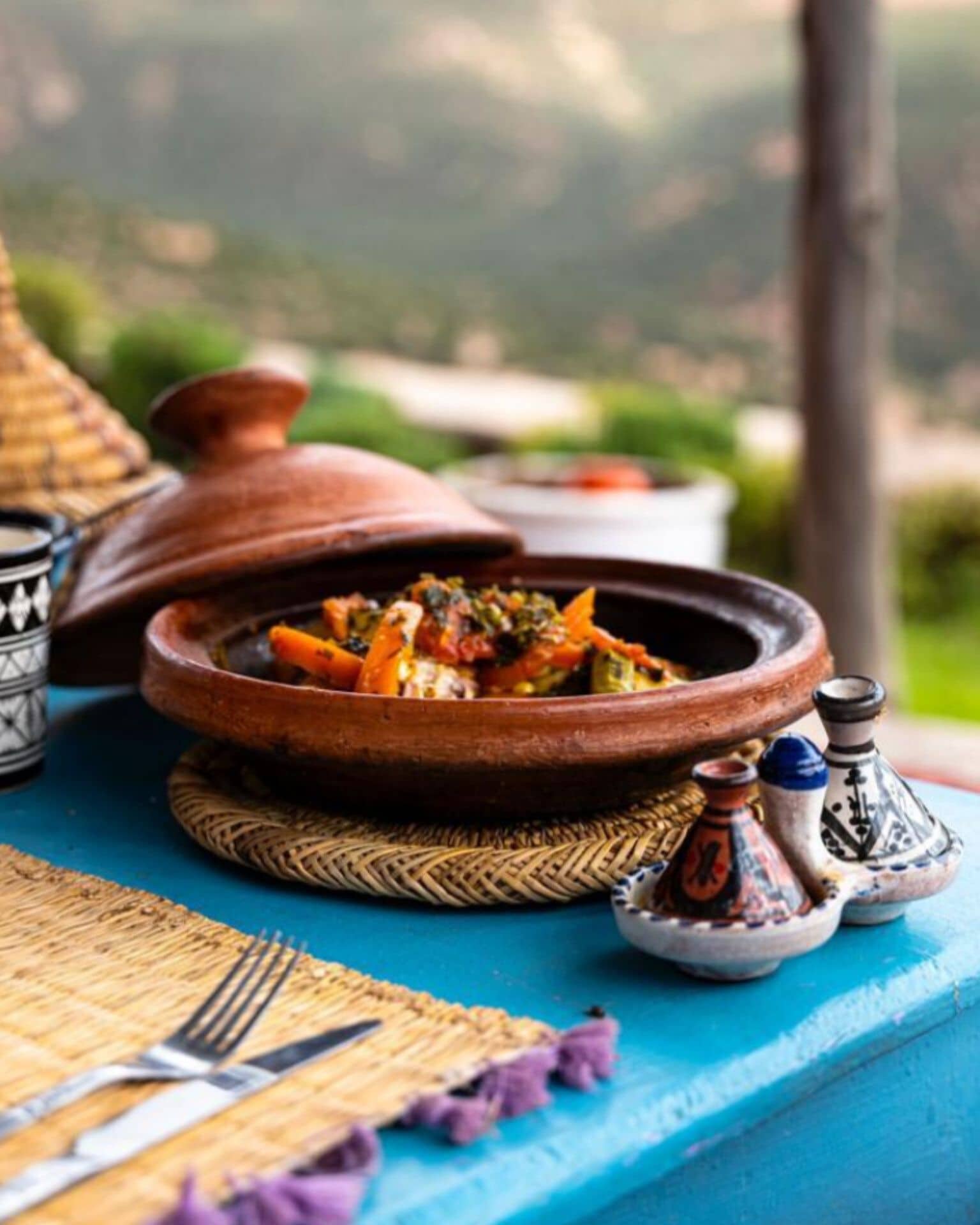 A traditional Moroccan tagine dish on a woven mat, with a view of greenery in the background.