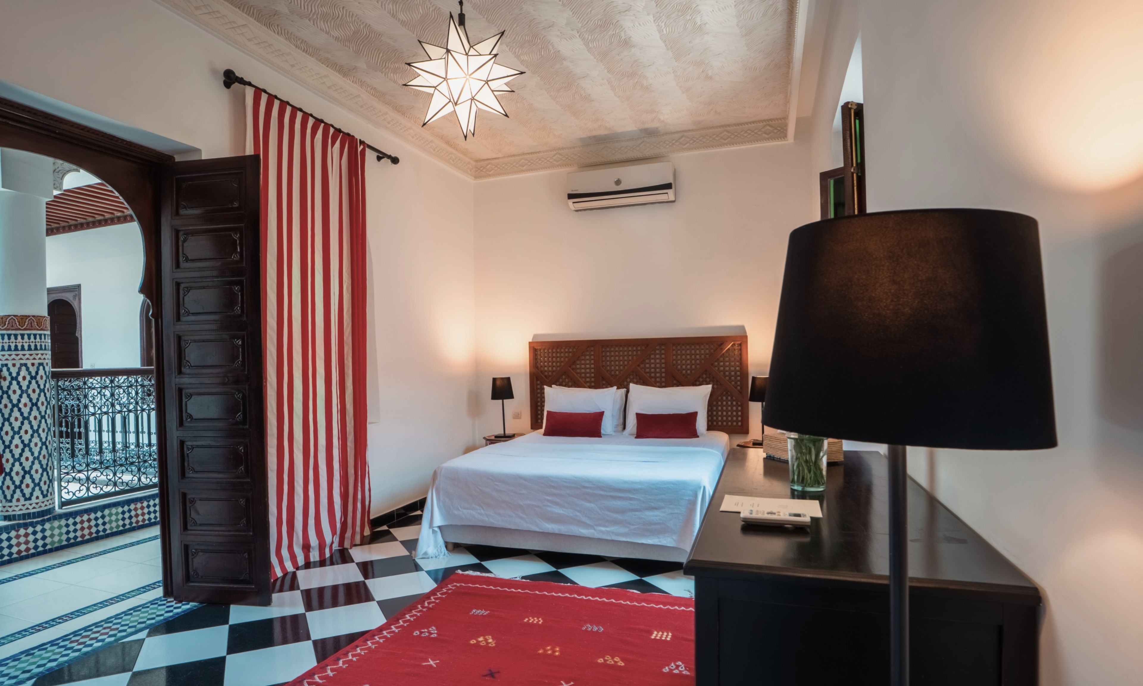 A hotel room with a checkered floor, a bed with white linens, a red and white striped curtain, a star-shaped ceiling light, and a wooden door.