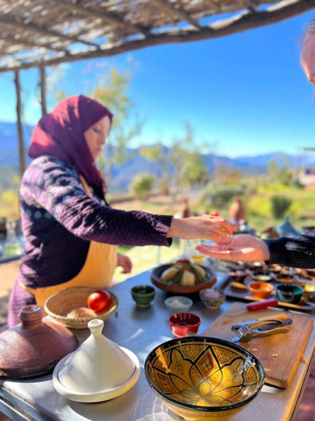 Uma pessoa servindo comida em uma mesa ao ar livre, com diversos pratos e utensílios.