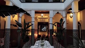 Un comedor con una larga mesa dispuesta para una comida, rodeado de columnas y arcos decorados, y un balcón en lo alto.