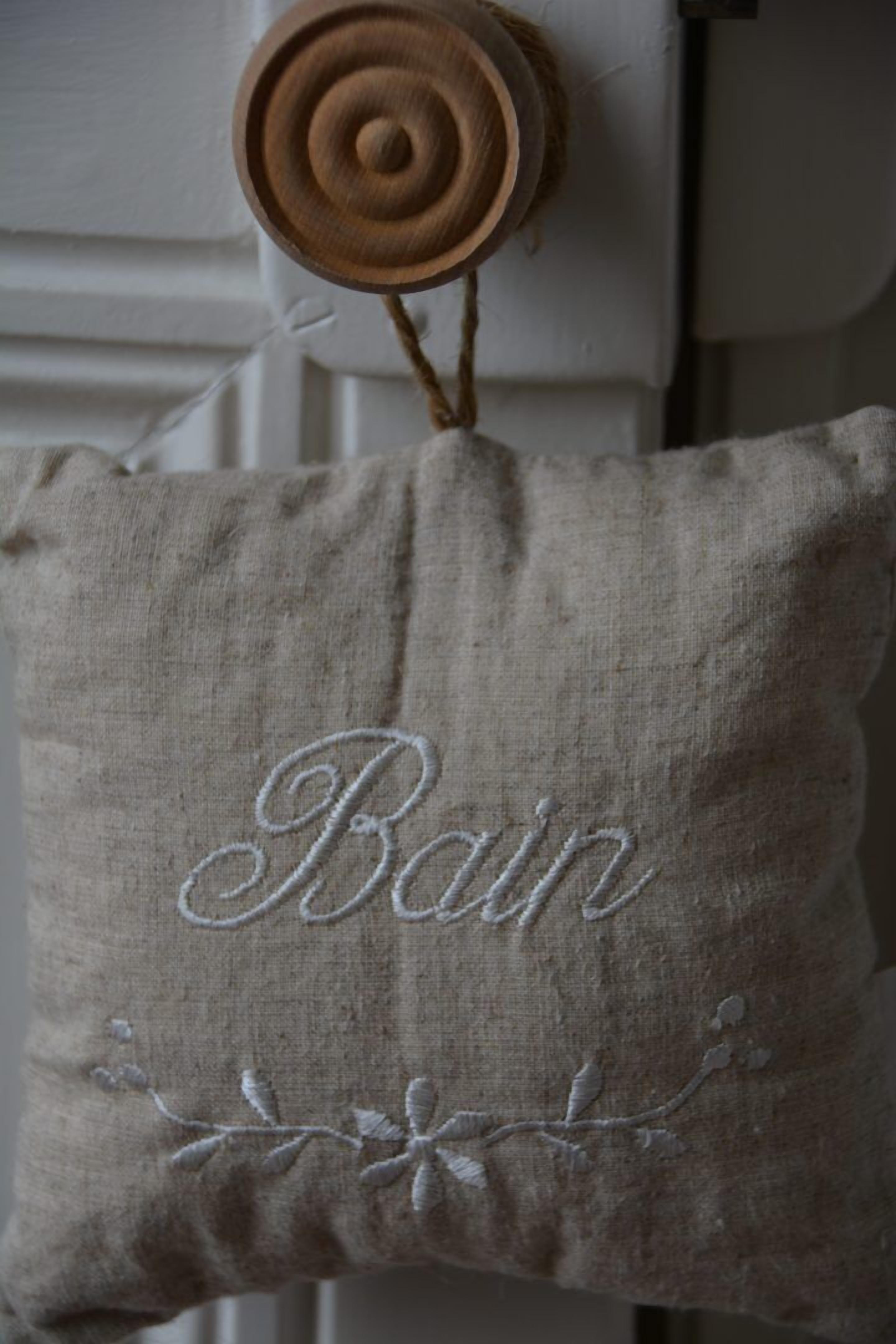 A beige pillow with embroidered text "Bain" and floral designs, hanging on a white wall.