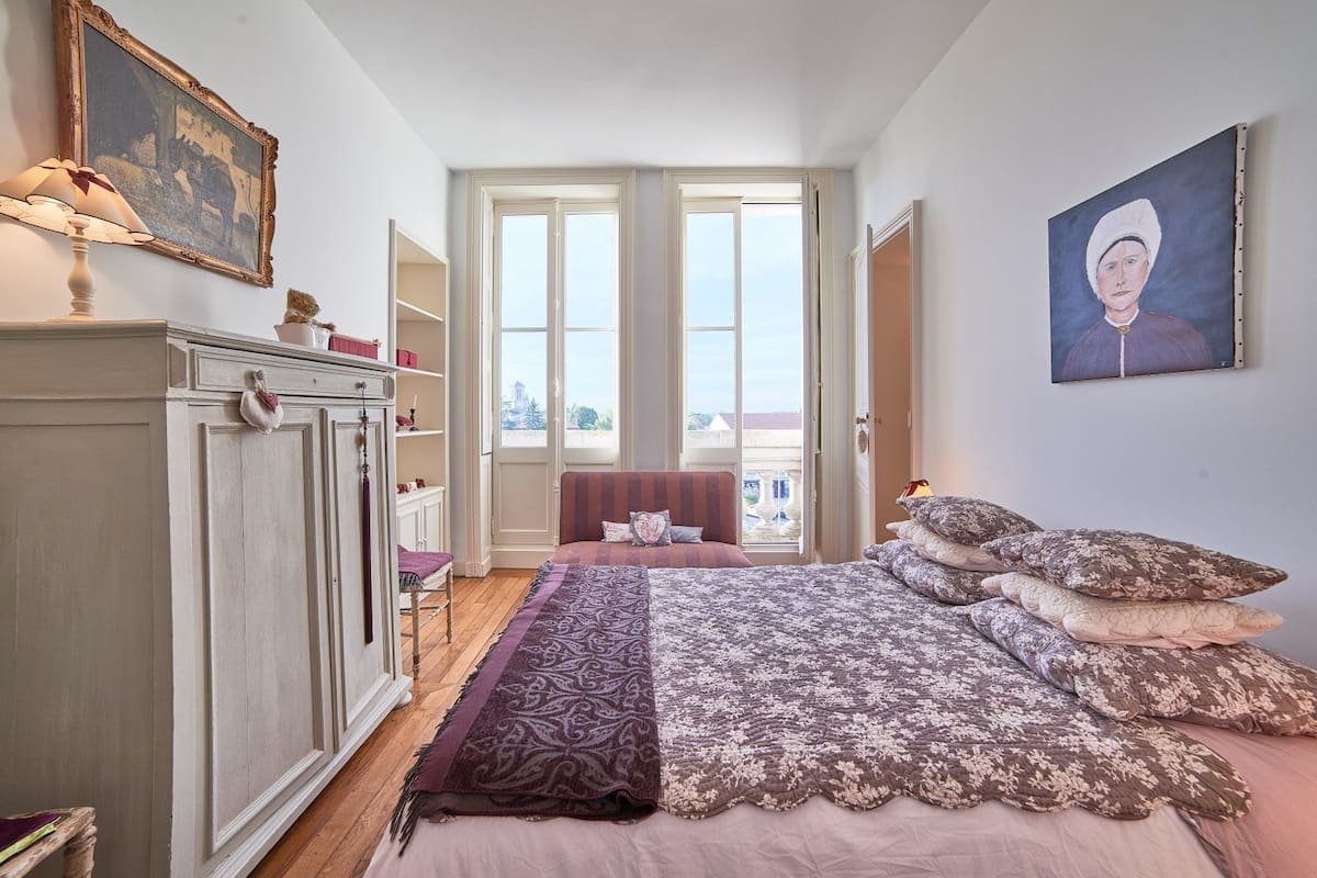 Een slaapkamer met een groot bed, een stoel, een dressoir en een raam.