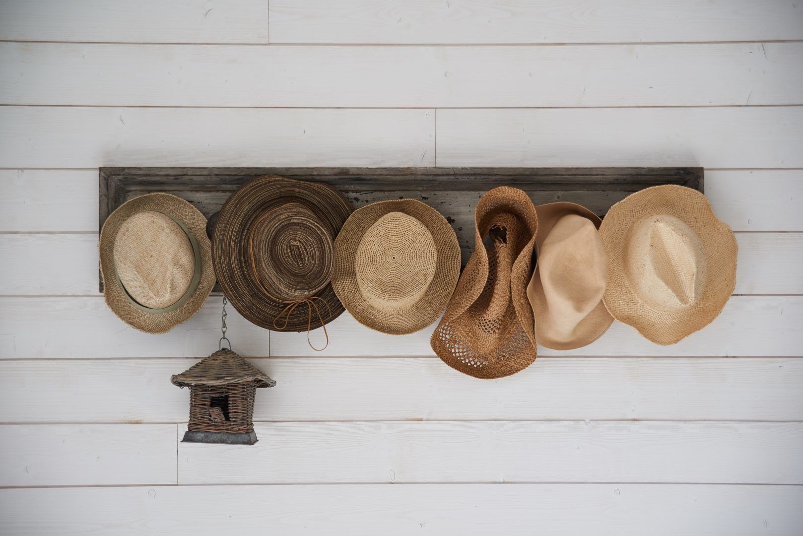 Una colección de sombreros tejidos y un pequeño farol de mimbre colgando de una pared de madera blanca.