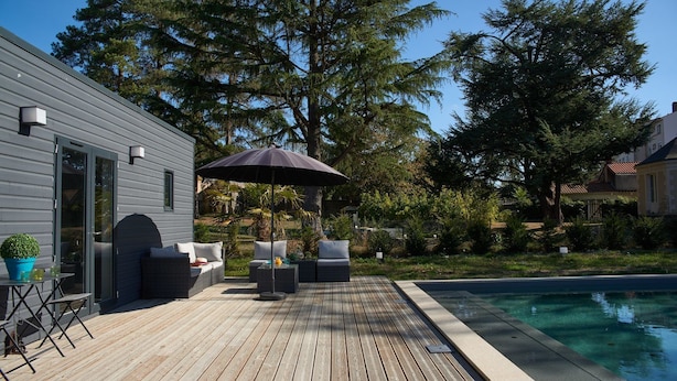 Una terraza de madera con mobiliario exterior, piscina y una casa de fachada gris.