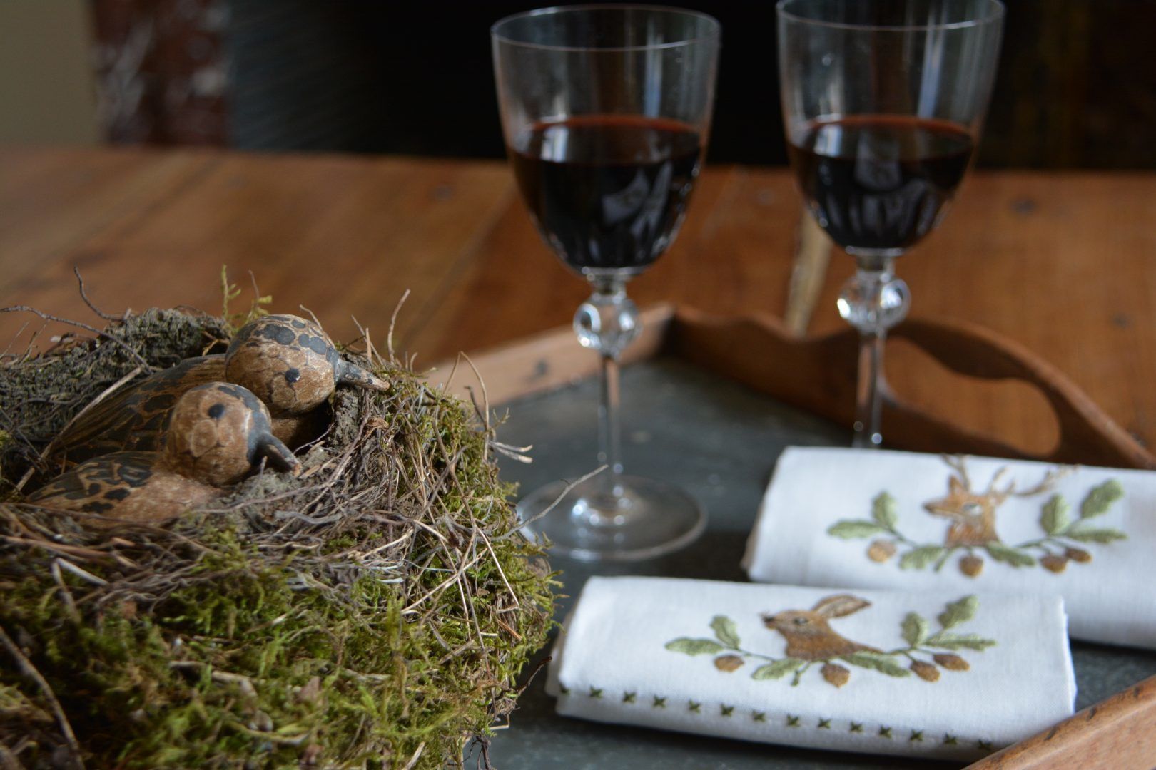Un nid contenant deux oisillons, deux verres à vin remplis d’un liquide rouge et un tissu orné d’un motif représentant un lapin.