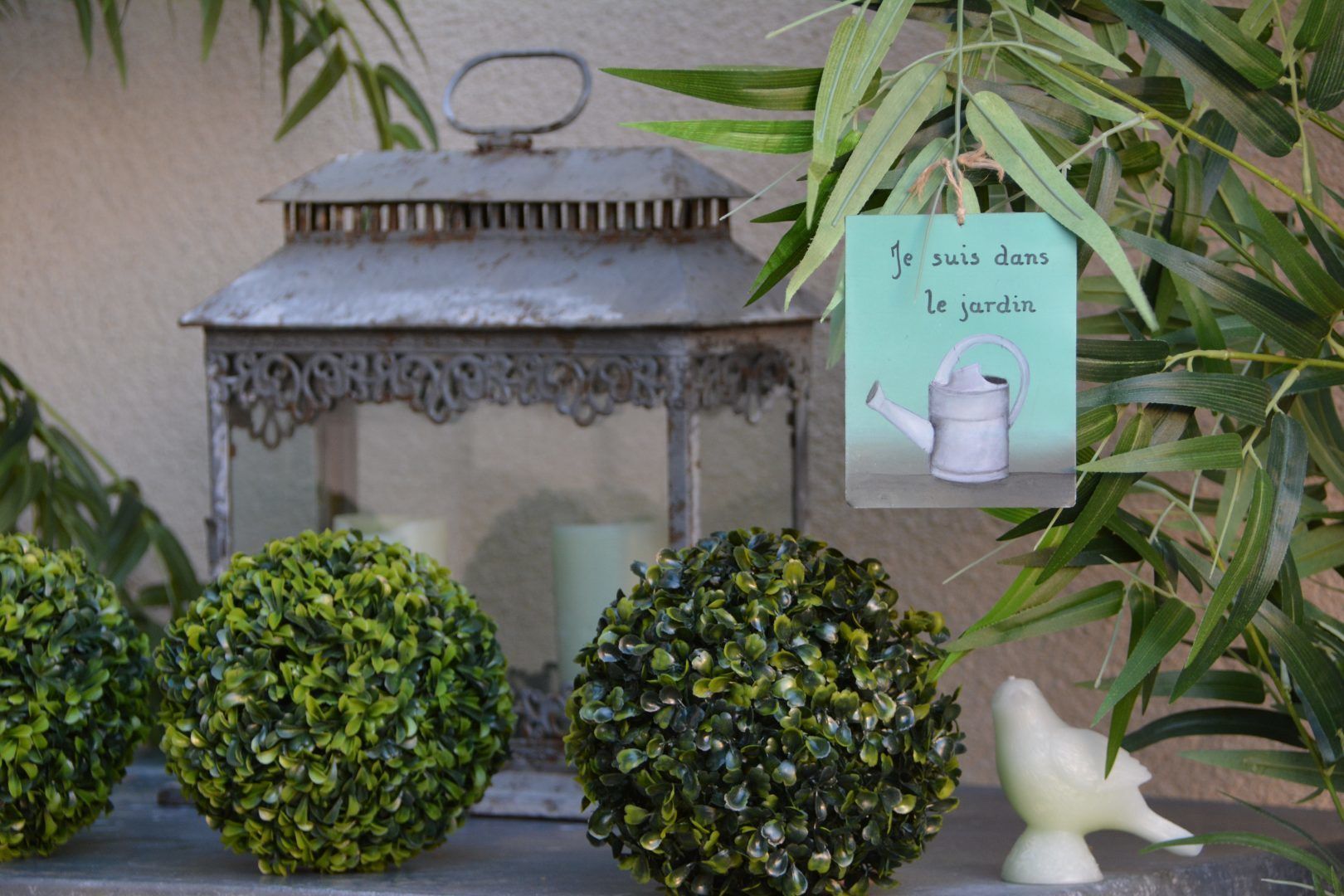 A decorative garden setting with a metal lantern, spherical bushes, and a small white bird statue.