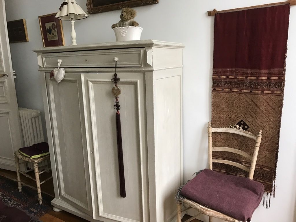 A white cabinet with a hanging ornament, a wooden chair with a purple cushion, and a patterned fabric draped over a chair.