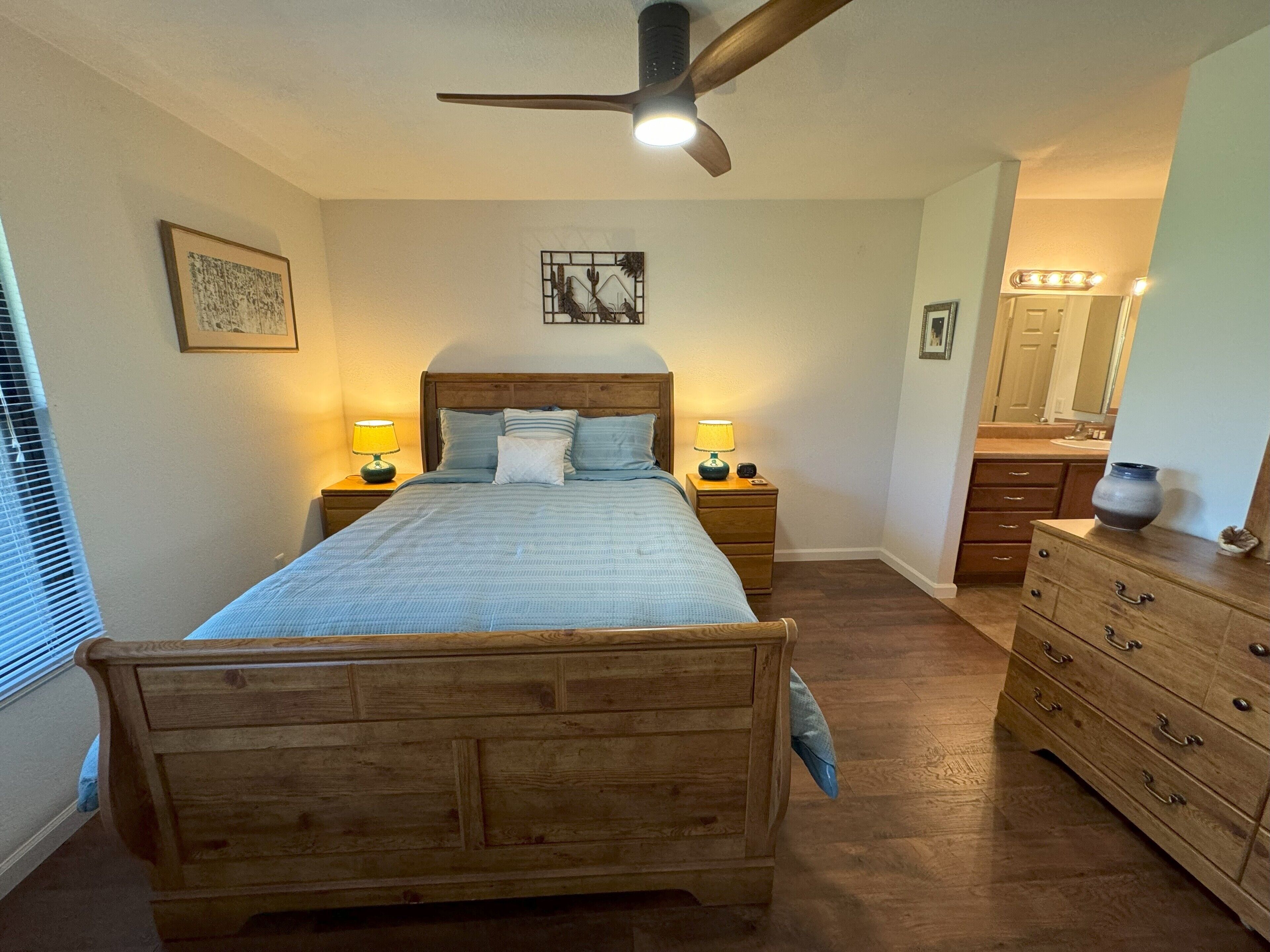 Une chambre à coucher avec un lit en bois, des lampes de chevet, un ventilateur de plafond, une commode et une salle de bain avec un meuble-lavabo et un miroir.