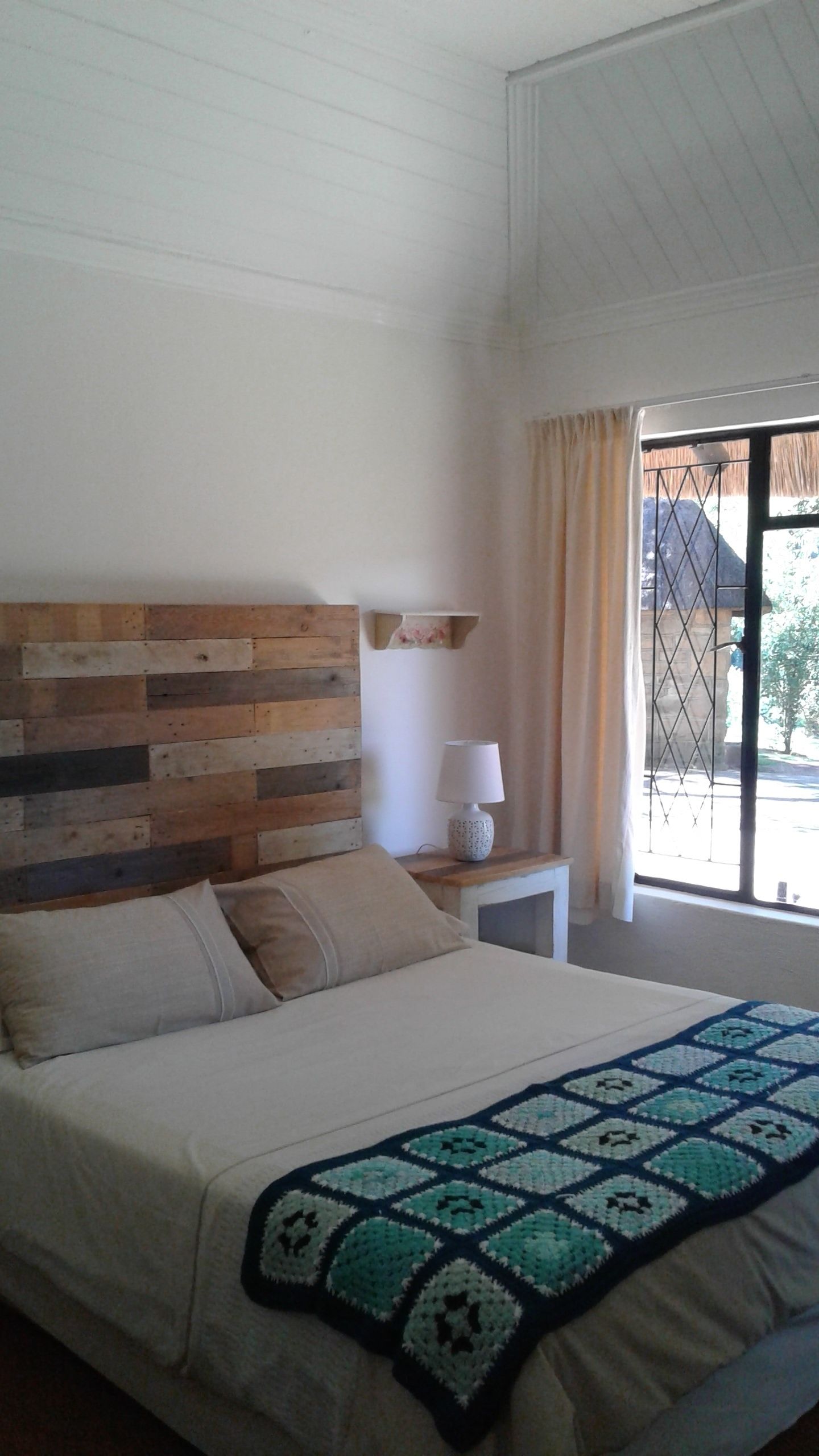 Un dormitorio con cabecera de madera, una mesita de noche con lámpara, una cama con colcha y vistas al exterior a través de una ventana.