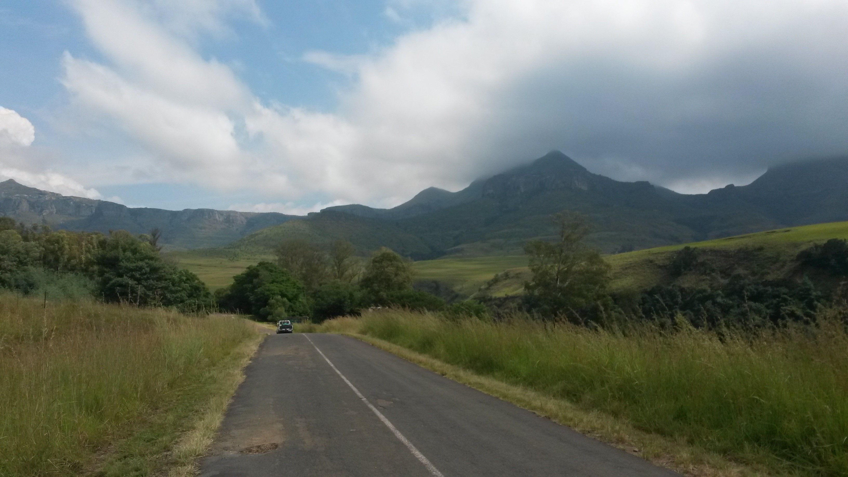 Una carretera rural bordeada por campos verdes y montañas bajo un cielo nublado.