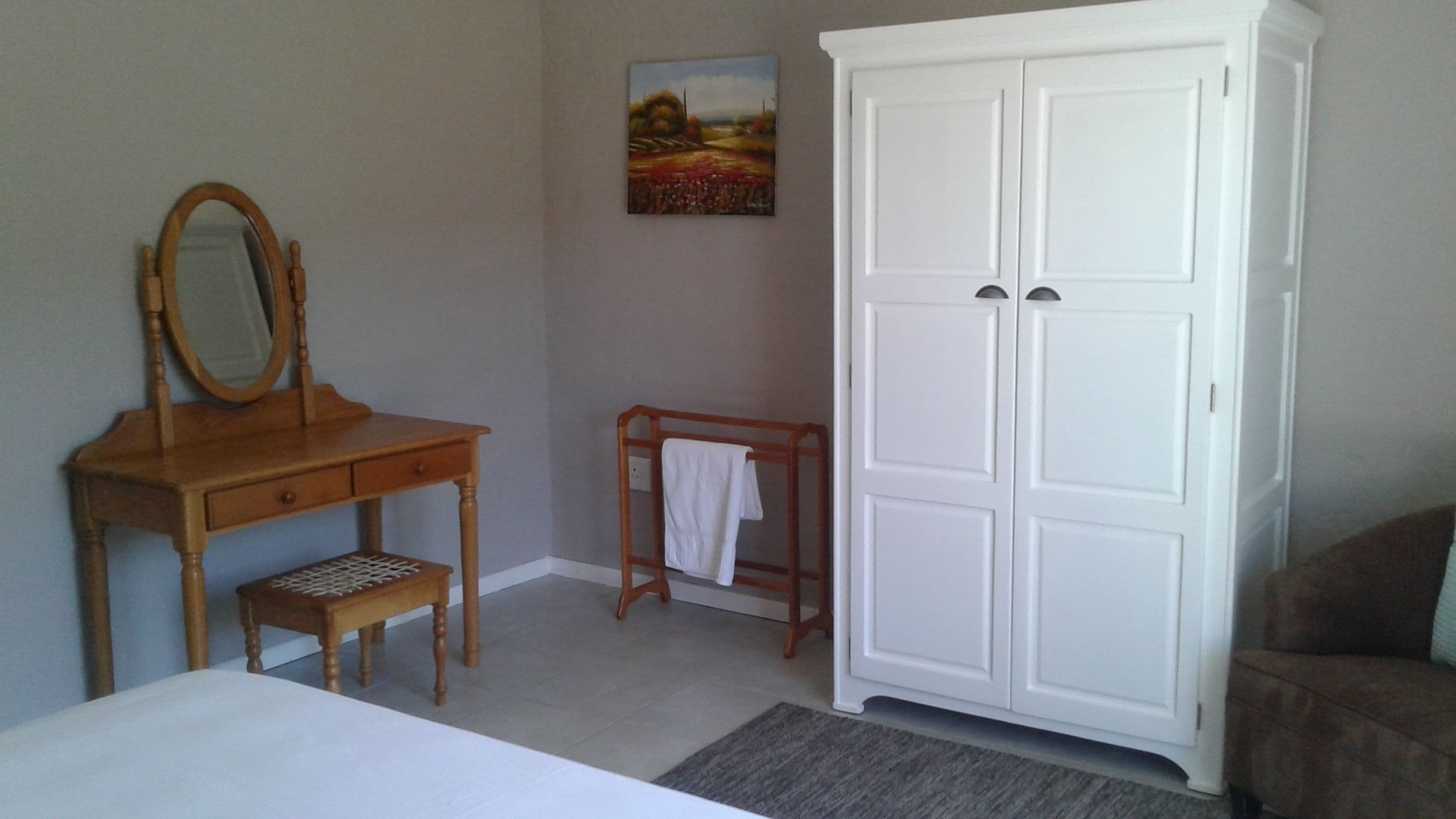 A bedroom with a wooden dresser, a mirror, a small wooden stool, a white wardrobe, and a painting on the wall.