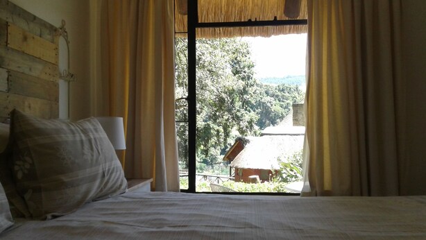 A bed with a pillow, a window with curtains, and a view of a building and trees outside. - Ledges Retreat (Jagersrust)