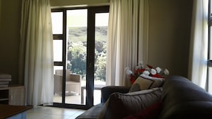 A living room with a sliding glass door leading to a balcony, a sofa, a small table, and a vase of flowers. - Ledges Retreat (Jagersrust)
