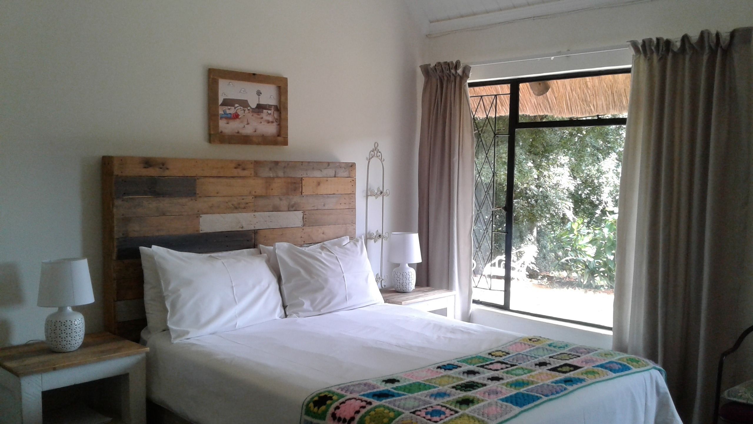 Quarto com cabeceira de madeira, roupa de cama branca, colcha colorida, abajur e janela com cortinas.