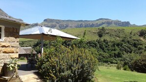 A stone building with a patio and a table under a metal umbrella, overlooking a lush valley with mountains in the background. - Ledges Retreat (Jagersrust)