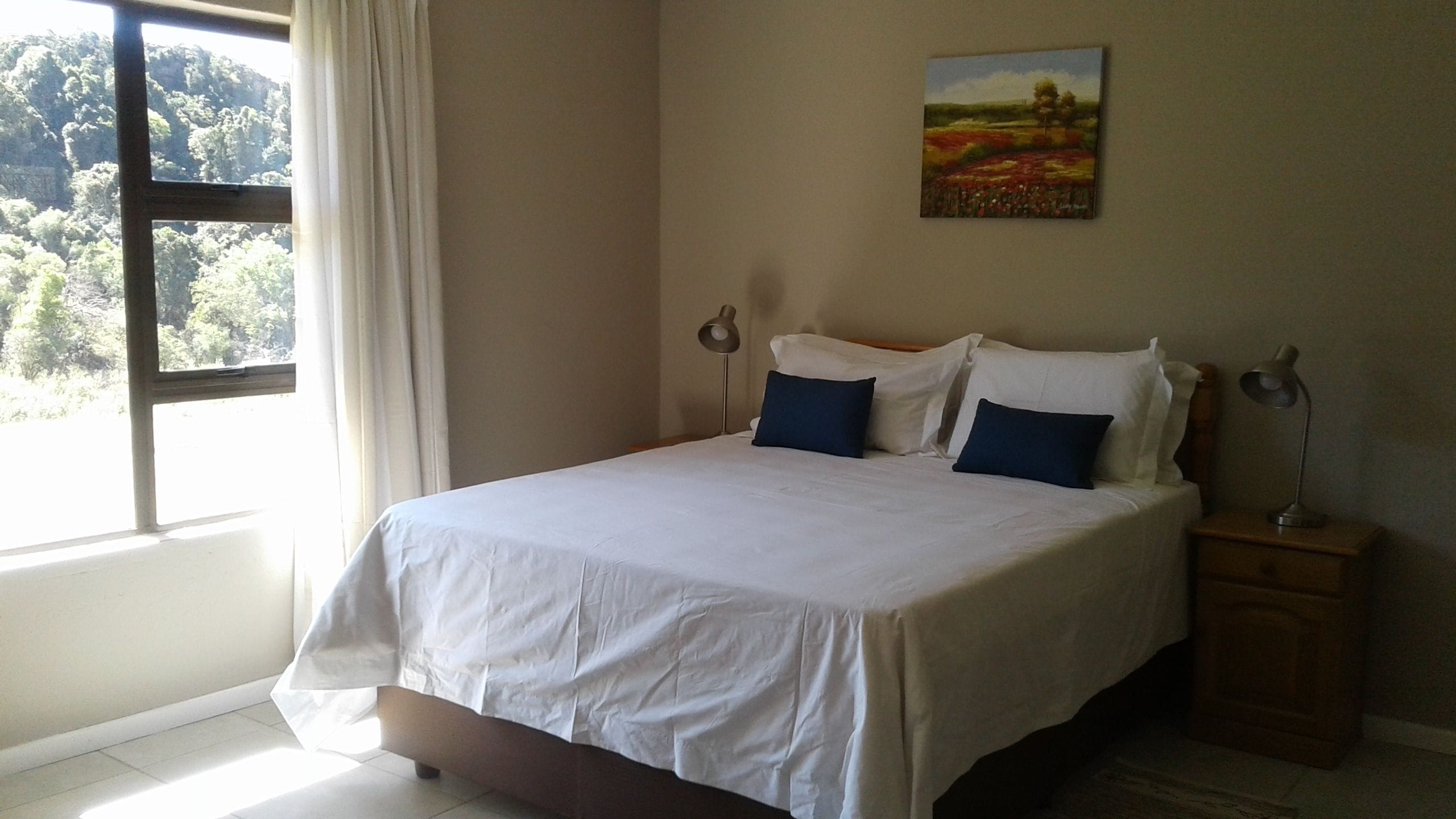 A neatly made bed with white bedding and two blue pillows, a bedside lamp, and a painting on the wall.