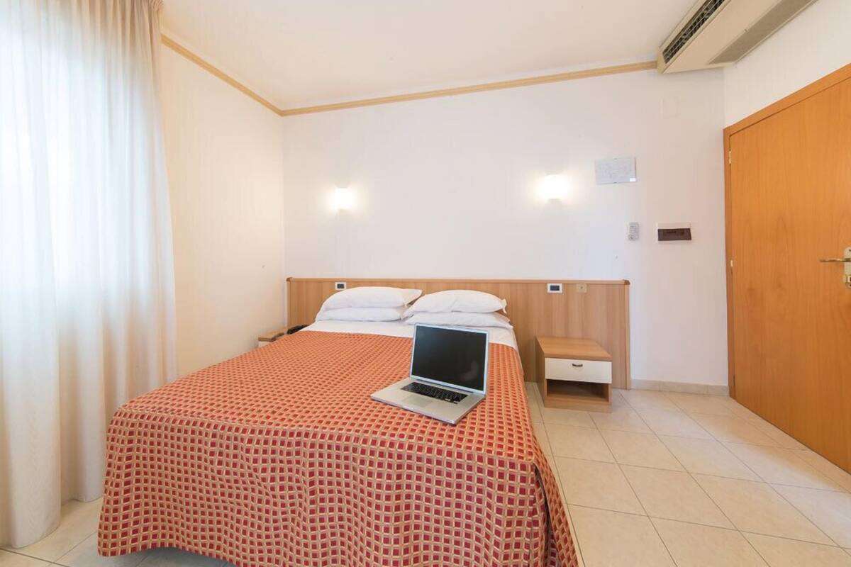 A hotel room with a bed, a laptop on the bed, a wooden headboard, a nightstand, a wooden door, and an air conditioning unit on the wall.