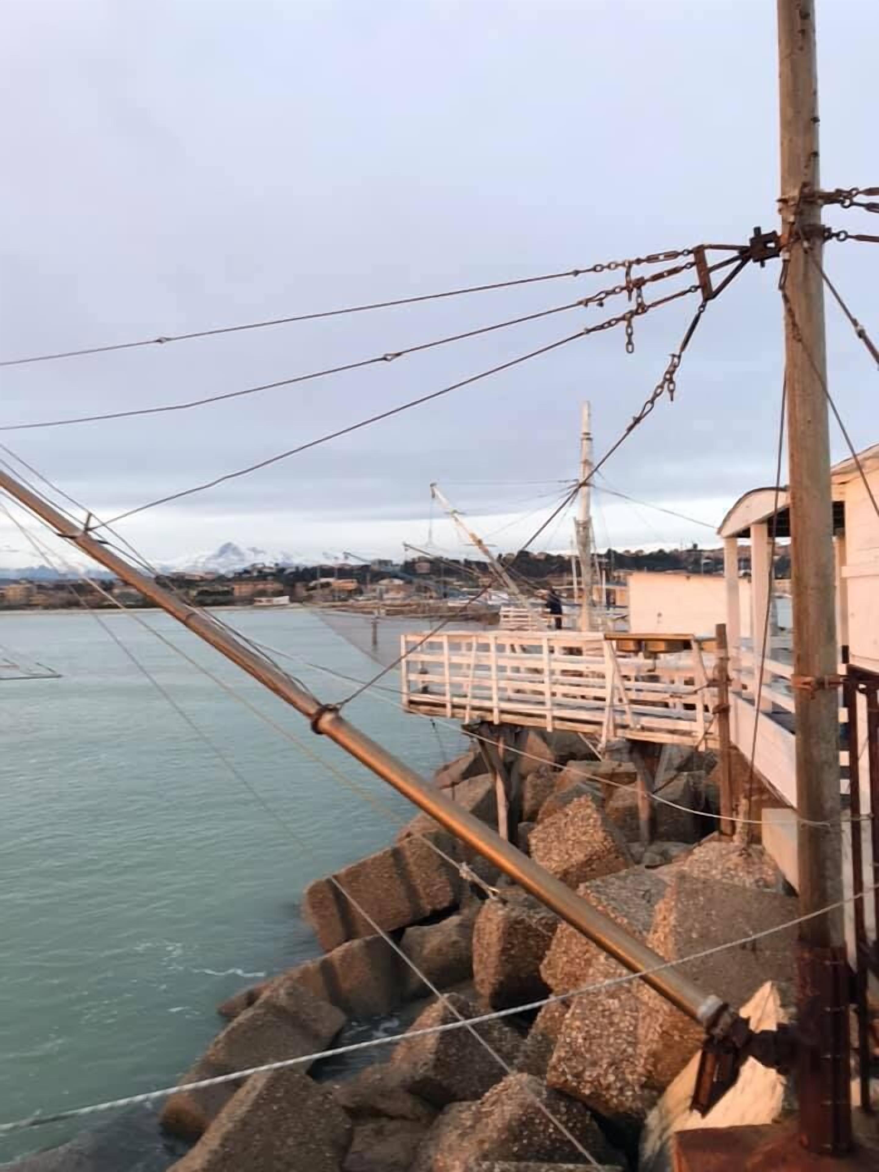 A coastal pier with a wooden walkway and a metal railing, overlooking calm waters and distant mountains.