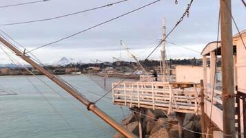 A coastal pier with a wooden walkway and a metal railing, overlooking calm waters and distant mountains.