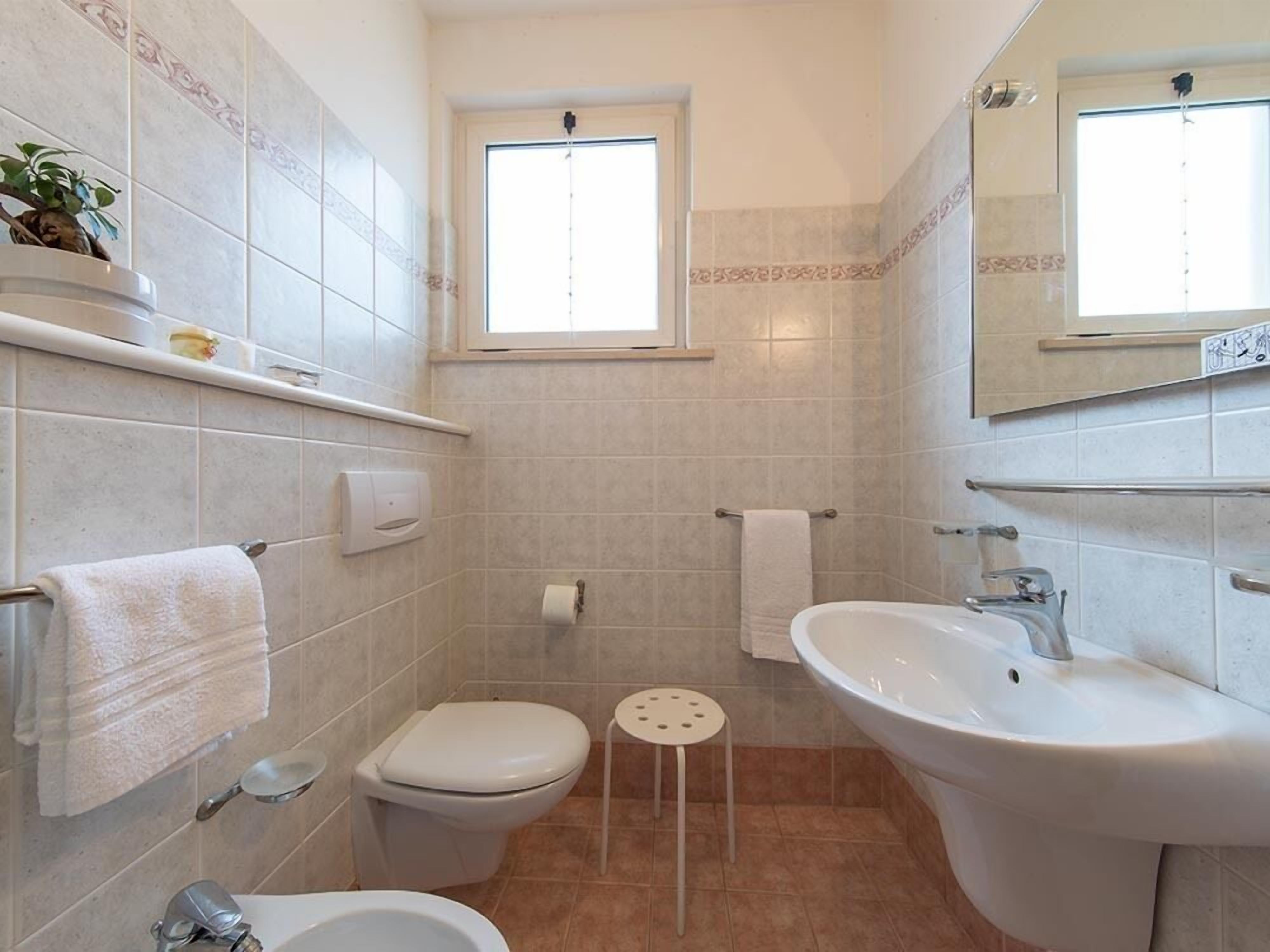 Un baño con inodoro, bidé, lavamanos y un taburete pequeño. Hay una ventana sobre el inodoro y una repisa con una planta y un objeto pequeño.