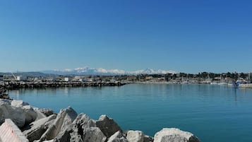A coastal marina with rocky breakwaters, boats, and a distant mountain range with snow-capped peaks.