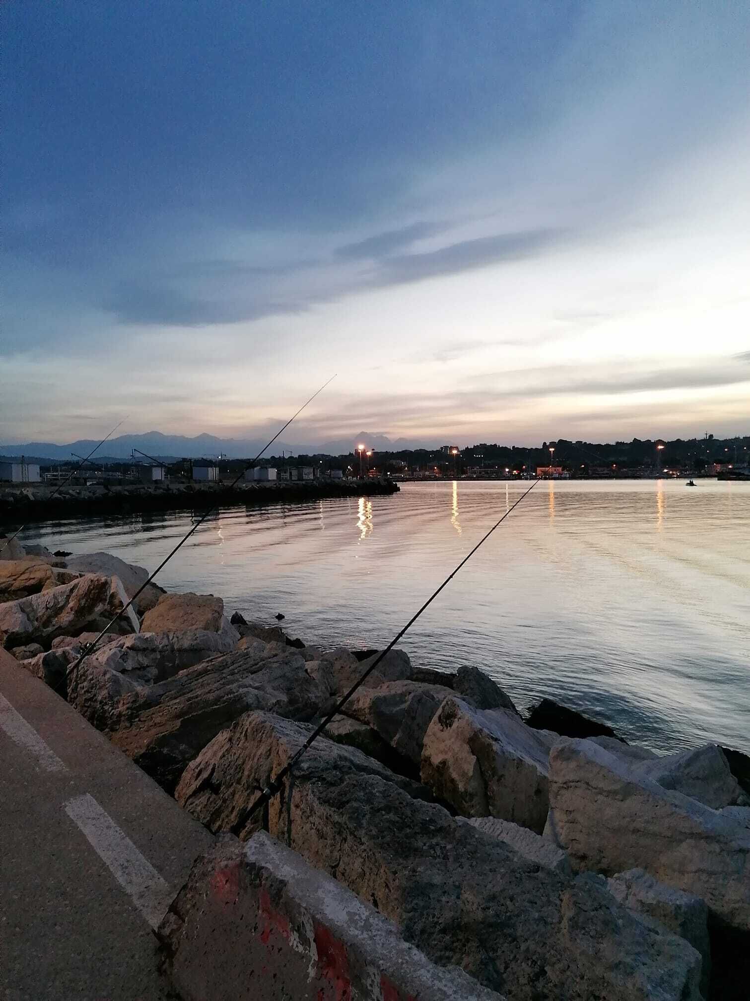 Um cais tranquilo com molinetes de pesca, pedras e luzes da cidade ao longe.