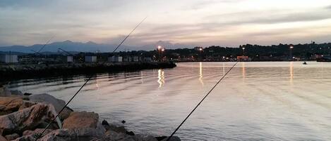 Um cais tranquilo com molinetes de pesca, pedras e luzes da cidade ao longe.