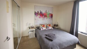 Ein Schlafzimmer mit Bett, Nachttisch, Spiegel, einem Fenster mit Vorhängen und einem Stadtbild an der Wand.