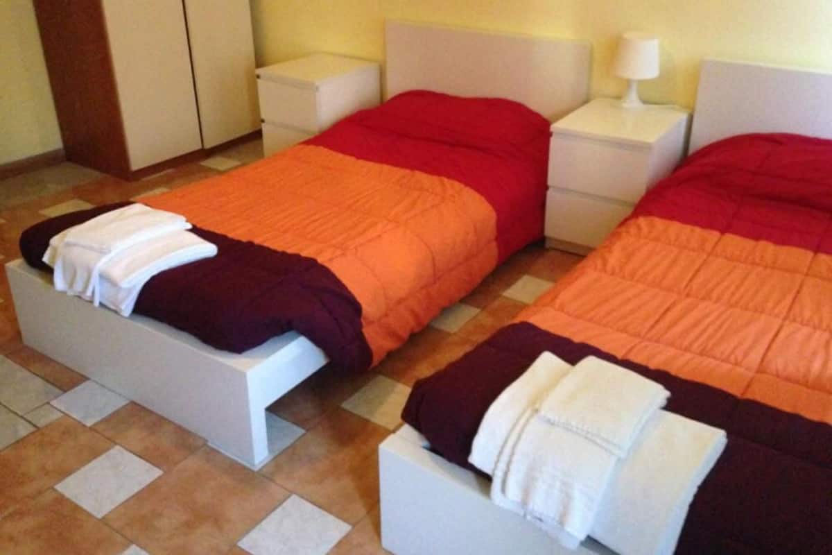 Una habitación de hotel con dos camas, cada una con cubrecama rojo y naranja, cabeceros blancos y mesitas de noche con lámparas.
