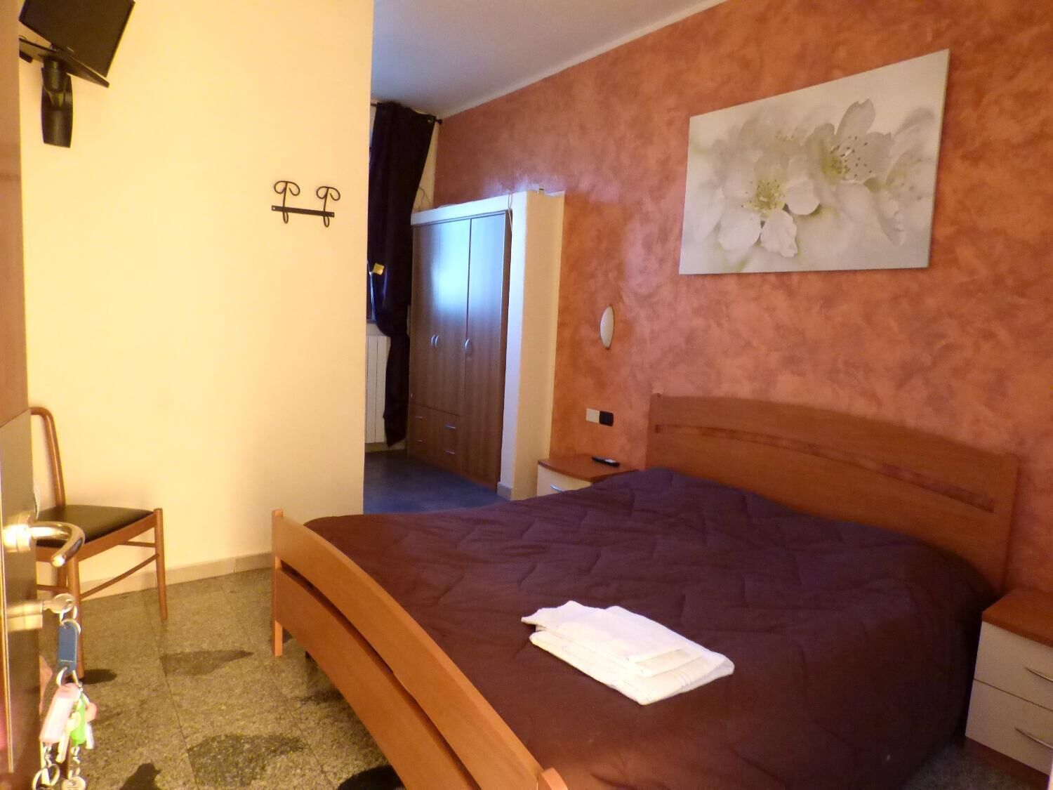 A bedroom with a wooden bed, a nightstand, a chair, a wardrobe, and a painting of flowers on the wall.