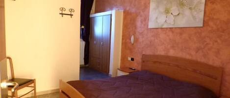 A bedroom with a wooden bed, a nightstand, a chair, a wardrobe, and a painting of flowers on the wall.