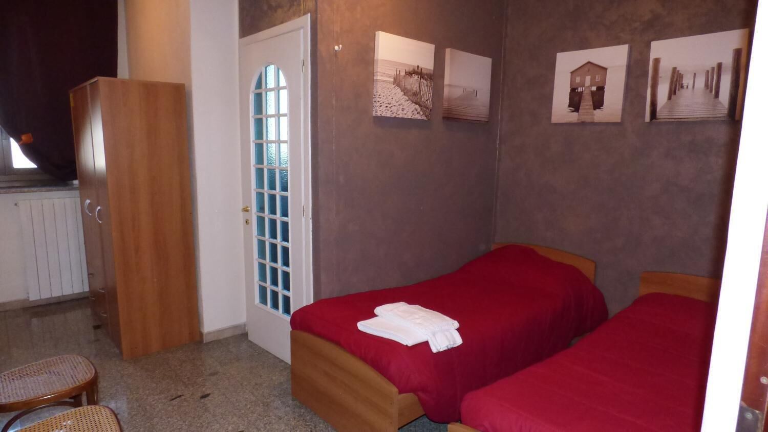 A bedroom with two single beds, a wooden wardrobe, a door with a window, and three framed pictures on the wall.