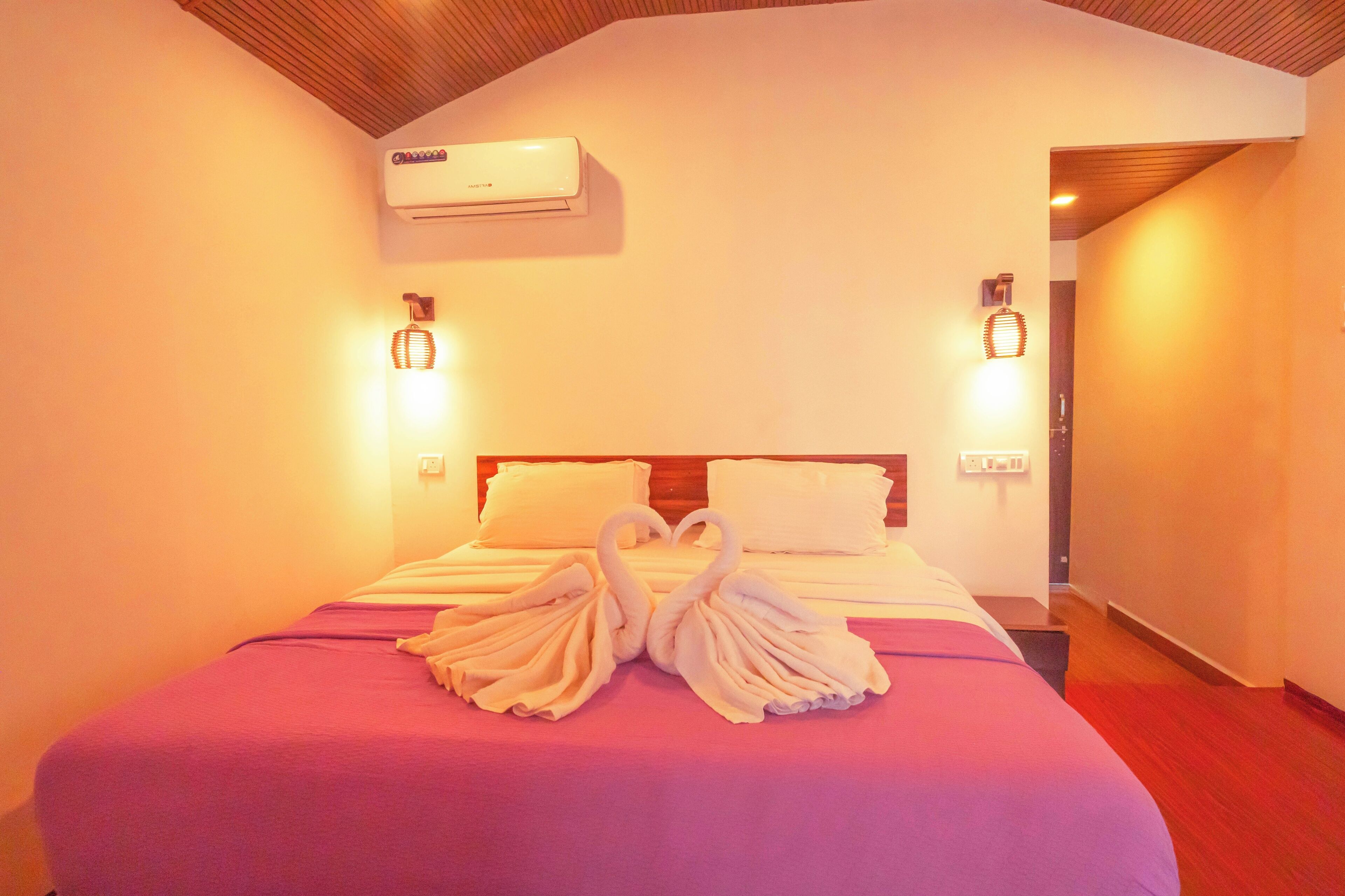 Habitación de hotel con una cama, dos lámparas fijadas a la pared, un aire acondicionado y un par de toallas dispuestas en forma de cisnes sobre la cama.