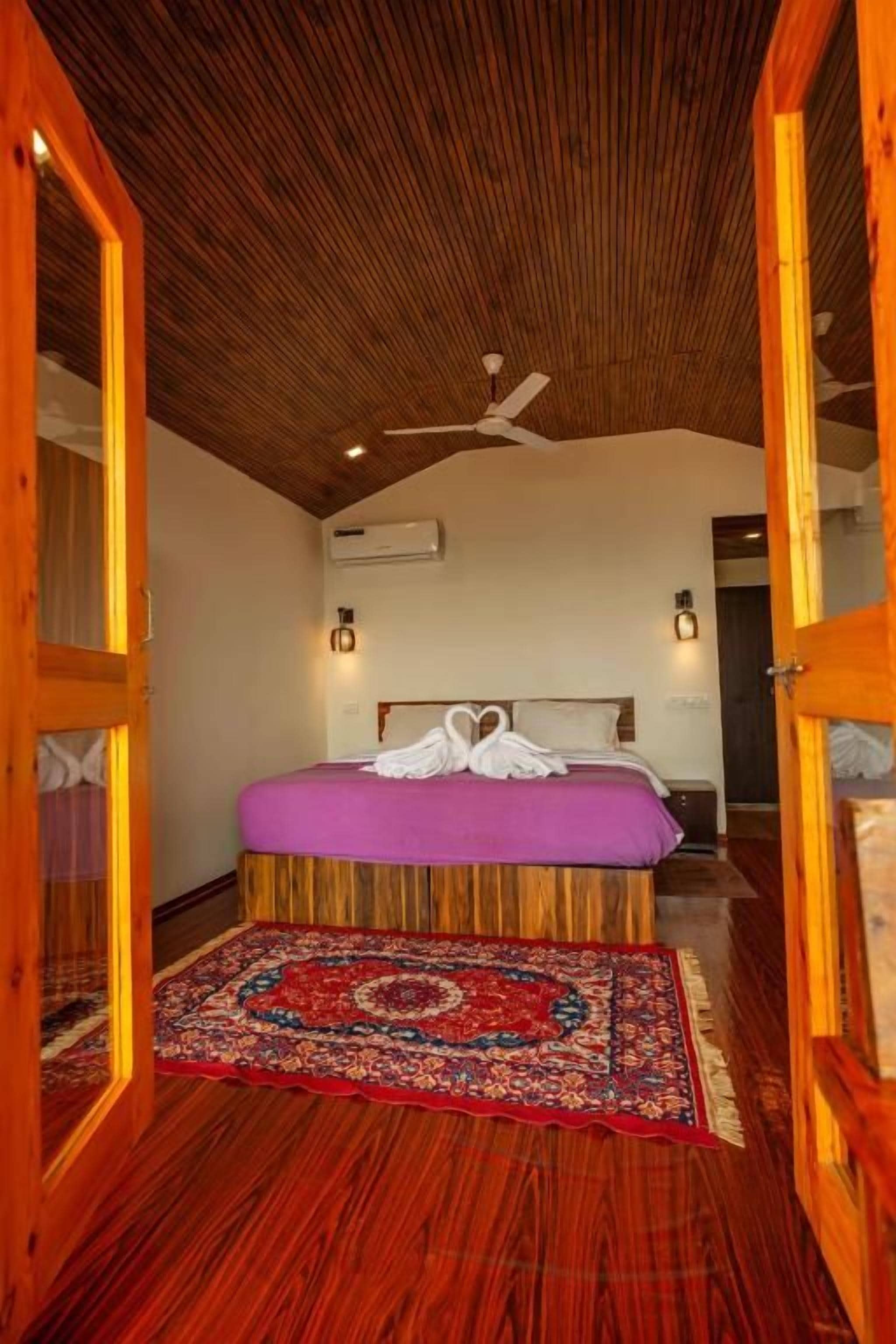A bedroom with a wooden ceiling, a bed with a purple bedspread, and a red patterned rug.