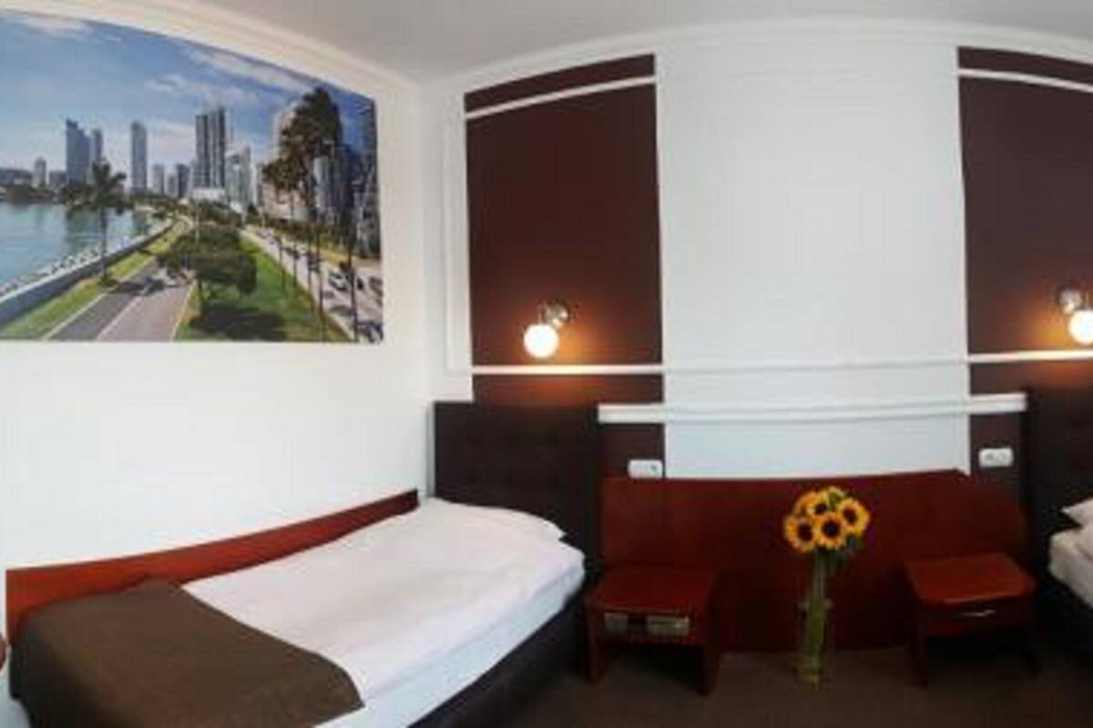 Ein Hotelzimmer mit zwei Einzelbetten, einem kleinen Tisch mit Blumenvase und einem Stadtbild an der Wand.
