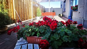 Une cour avec une clôture en bois, un banc, des pots de fleurs rouges et un parasol blanc orné d’un logo.
