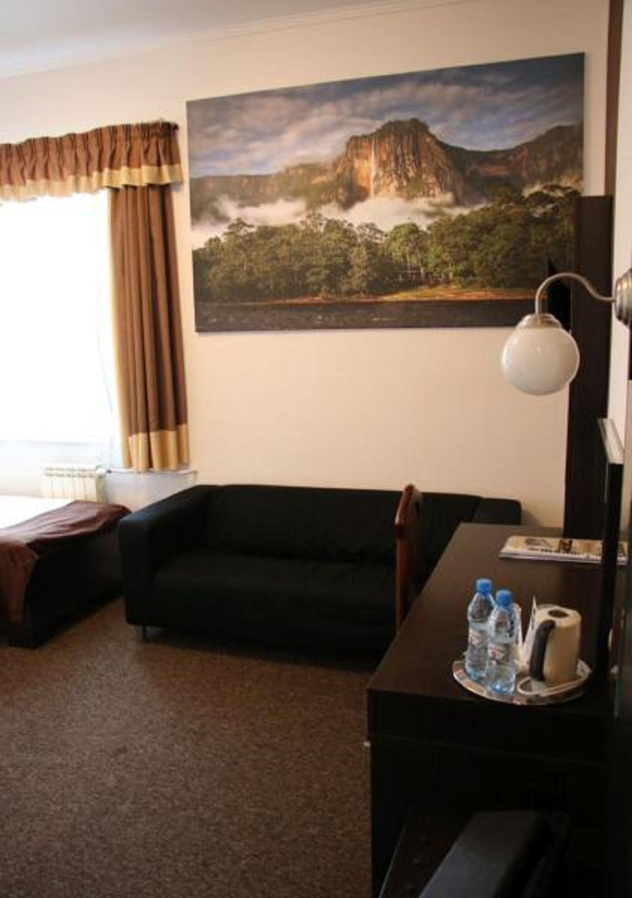 Una habitación de hotel con un sofá negro, un escritorio con lámpara, un cuadro de un paisaje montañoso y una ventana con cortinas.