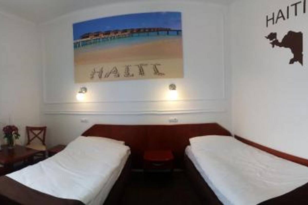 Ein Hotelzimmer mit zwei Einzelbetten, einem Schreibtisch mit Stuhl, einem Fenster mit Vorhängen sowie einem Wandbild mit Strandmotiv und dem Schriftzug "HAITI".